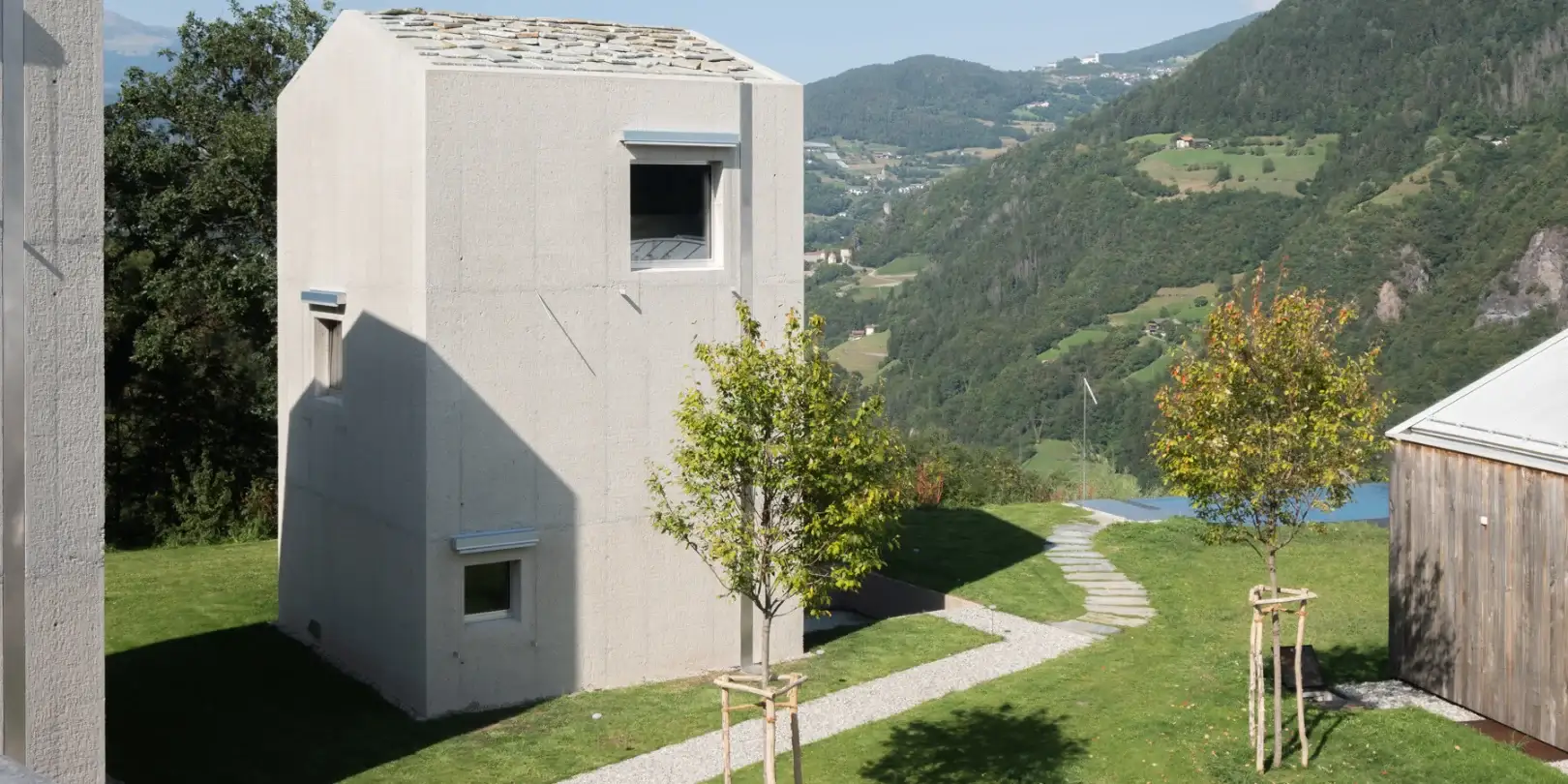 Moderne Garten-Neuanlage für ein Gebäude mit Betonarchitektur in Hanglage mit gepflegtem Rasen, gepflastertem Weg und angelegten Jungbäumen im Grünen.